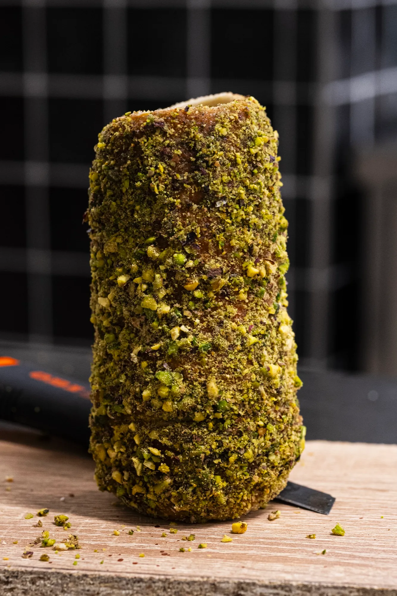 A chimney cake coated in green crushed pistachios on a wooden board